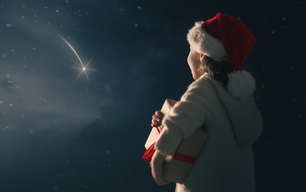 Merry Christmas Cute little child with xmas present. Happy kid is making wish looking at falling star. Portrait of girl with gift on dark sky background